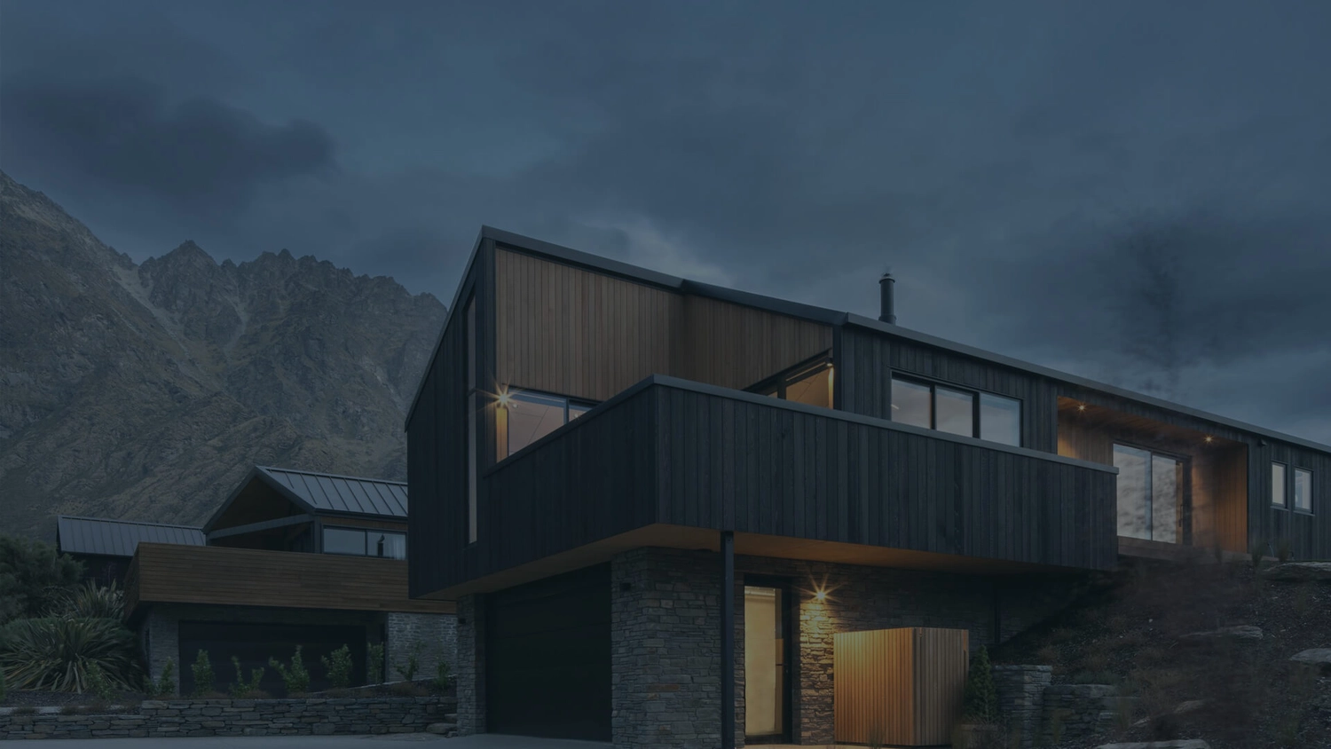 GM-Fensterbau - Fenster - Türen - Rollladen - Ein modernes, dunkel verkleidetes Architekturhaus mit Holzakzenten und Steinfundamenten liegt unter einem stimmungsvollen, bedeckten Abendhimmel vor einer schroffen Bergkulisse.
