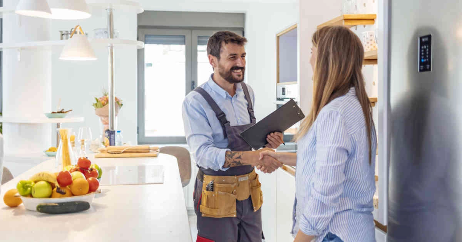 GM-Fensterbau - Fenster - Türen - Rollladen - Ein lächelnder Techniker schüttelt einer Frau in einer Küche die Hand, während er ein Klemmbrett in der Hand hält.