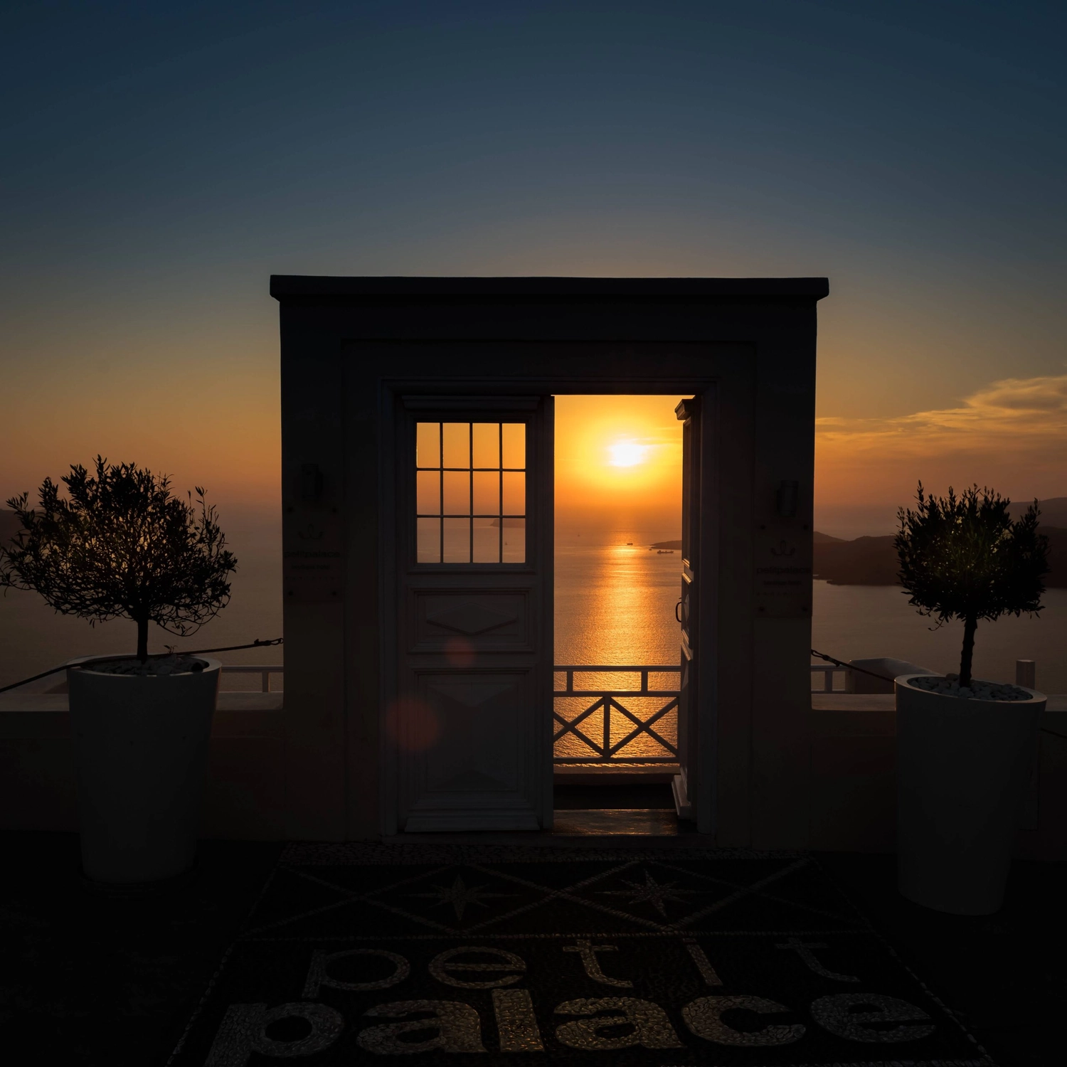 GM-Fensterbau - Fenster - Türen - Rollladen - Eine steinerne Tür öffnet sich zu einem lebendigen Sonnenuntergang über dem Meer über Santorini und umrahmt die Spiegelung der Sonne auf dem Wasser.