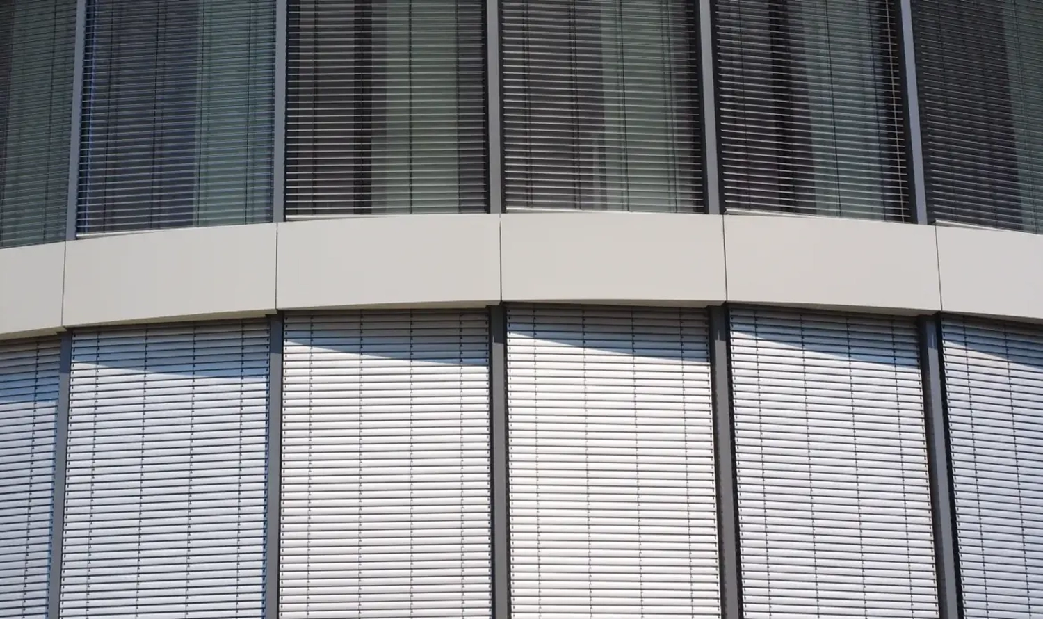 GM-Fensterbau - Fenster - Türen - Rollladen - Eine geschwungene Gebäudefassade mit großen Glasfenstern, die von horizontalen grauen Jalousien und außenliegenden Rollläden verdeckt werden.